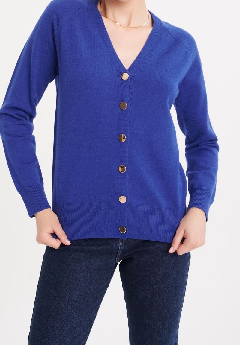 Femme portant un cardigan bleu roi à col en V avec sept boutons dorés, associé à un jean bleu foncé, les mains reposant près des poches.
