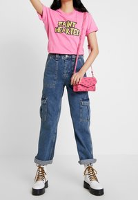 Camiseta gráfica rosa con texto "FAINT HEARTED", pantalones cargo de mezclilla azul de tiro alto y botas blancas con plataforma y cordones. Pequeño bolso rosa cruzado.