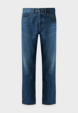 Jeune homme portant un pull en maille marron zippé, une chemise beige clair, un jean bleu et des chaussures en cuir marron, debout devant un fond blanc uni.
