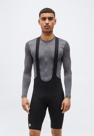 Muž oblečený v sivom dlhom cyklistickom drese s dlhým rukávom a čiernych trenírkach s trakmi stojaci na bielom pozadí.