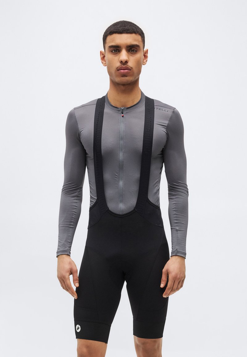 Άνδρας που φορά γκρι μακρυμάνικη μπλούζα ποδηλασίας και μαύρο σορτς με τιράντες, στέκεται μπροστά σε απλό λευκό φόντο.