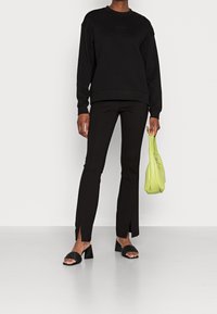 Černá mikina s kulatým výstřihem, spárovaná s form-fitting černými kalhotami s bočními rozparky. Doplňky zahrnují svěže zelenou tašku a černé pantofle.