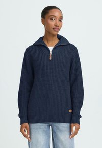 Marineblauwe gebreide pullover met een half-rits kraag, textuurstof, geribde manchetten en onderrand, voorzien van een kleine bruine logo patch aan de zijkant.