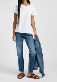 Camiseta blanca de algodón de manga corta, vaqueros rectos azules, sandalias rojas y marrones, sosteniendo una chaqueta de mezclilla. Atuendo simple y casual.