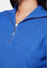 Pull en tricot côtelé bleu avec un col zippé, présentant un tissu texturé et un détail de fermeture éclair en argent.