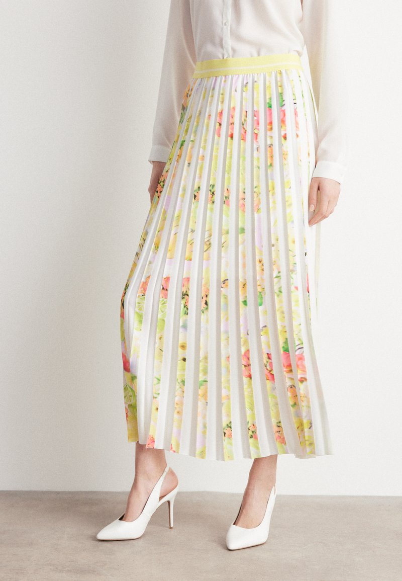 Frau, die einen plissierten Midikleid mit gelbem Blumenmuster, eine weiße Bluse und weiße Slingback-High-Heels trägt, steht drinnen auf einem beigen Boden.