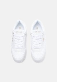 Sneakers bianchi Guess con lacci, motivo trapuntato e dettagli del logo dorato, visti dall'alto su uno sfondo semplice.