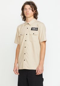 Kortærmet, beige skjorte med knapper lavet af letvægtsstof, med en brystlomme med et sort logo-patch og kontrasterende sorte knapper.