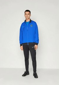 Blaue Zip-Jacke mit schwarzen Akzenten und Logo, über einem blauen Oberteil getragen, kombiniert mit schwarzen Hosen mit einem weißen Logo und schwarzen Schuhen.