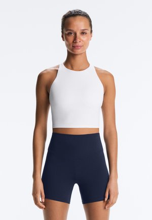 Kvinde iført en hvid, ærmeløs crop top og marineblå cykelshorts med høj talje, stående vendt mod fronten på en lys baggrund.