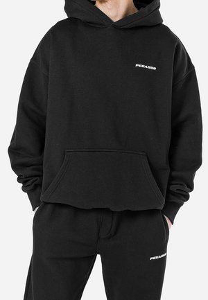 Hoodie noir surdimensionné avec une poche frontale, arborant un logo blanc "PEGADOR" sur la poitrine. Le matériau est doux, avec une texture semblable à de la polaire.