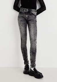 Jean skinny taille haute noirs avec un délavage gris, présentant cinq poches et des surpiqûres latérales contrastées. Associés à des mocassins noirs surdimensionnés.