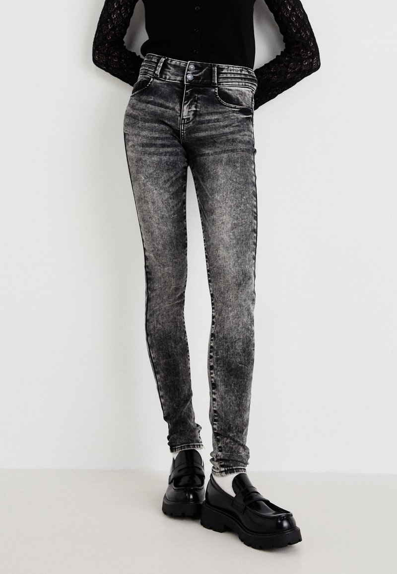 Jean skinny taille haute noirs avec un délavage gris, présentant cinq poches et des surpiqûres latérales contrastées. Associés à des mocassins noirs surdimensionnés.