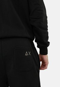 Pantaloni jogger neri con una tasca a toppa che presenta un design del logo triangolare. Il materiale sembra morbido con una finitura liscia e testurizzata.
