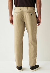 Pantalons en lin beige à coupe droite, avec deux poches arrière et une texture lisse, associés à des chaussures décontractées marron foncé.