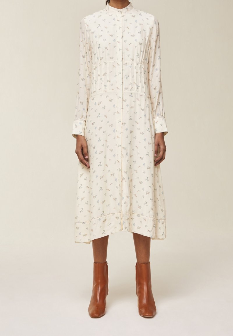 Kvinna står klädd i en krämfärgad midiklänning med små blommönster och bruna anklar stövlar, neutral bakgrund.