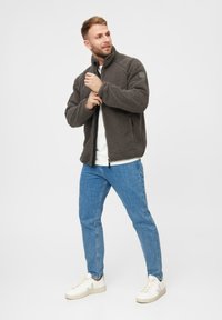 Giacca in pile grigia scura con colletto alto, zip completa, tessuto strutturato, abbinata a jeans azzurri chiari e sneakers bianche.
