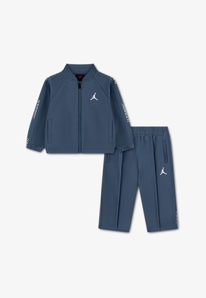 Tamsiai mėlynas Jordan sportinis kostiumas, kuriame yra striukė su užtrauktuku ir kišenėmis bei derančios kelnės. Abu drabužiai papuošti baltomis logotipo detalėmis.
