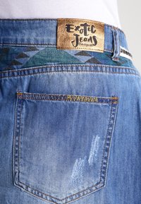 Jean en denim de couleur bleu délavé, avec une étiquette dorée, une ceinture à motifs géométriques et des coutures jaunes contrastées sur la poche arrière.