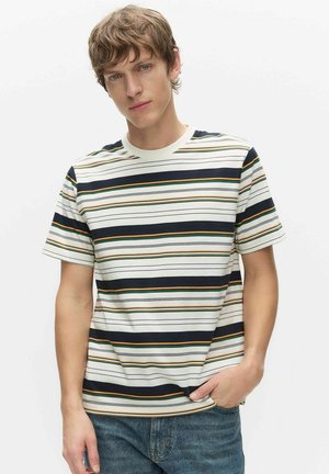 Ung man med kort brunt hår som bär en vit, marinblå, grön och orange randig t-shirt och blå jeans, med handen i fickan.