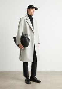 Abrigo de lana de color gris claro con un diseño de doble botonadura, llevado sobre un suéter negro de cuello alto. Un gorro negro, un bolso negro y zapatos completan el look.