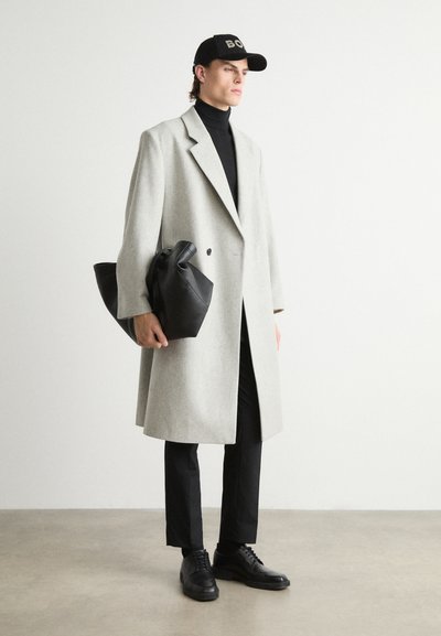 Manteau en laine gris clair avec un design à double boutonnage, porté sur un col roulé noir. Une casquette noire, un sac noir et des chaussures complètent le look.