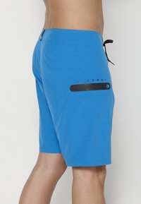 Bañador azul con cinturilla ajustable, bolsillo lateral con un acento negro y el logo "KOROSHI" impreso encima del bolsillo.