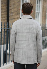 Harmaa ruudullinen takki, jossa on lävistetty kaulus, teksturoitu kangas ja rento malli, nähtynä takaa tiiliseinän taustalla.