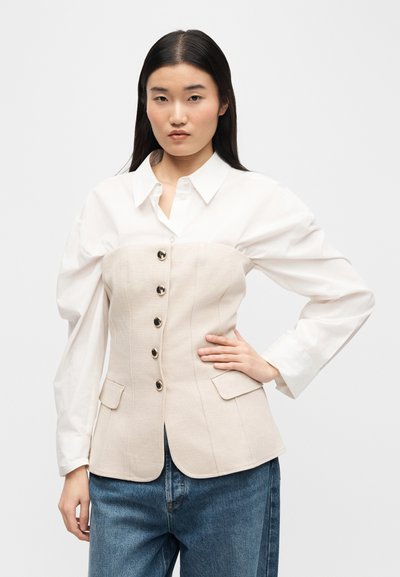 Νεαρή γυναίκα με μακριά σκούρα μαλλιά, φορώντας λευκό πουκάμισο με κουμπιά και ένα μπεζ γιλέκο σε στυλ κορσέ, συνδυασμένο με μπλε τζιν, ποζάρει με το χέρι στη μέση.