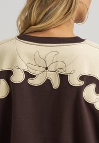 Γυναίκα με ξανθά μαλλιά που φορά μπλούζα σε μπεζ και σκούρο καφέ χρώμα, με κεντημένα λουλούδια και σπειροειδή μοτίβα στον επάνω μέρος της πλάτης και στους ώμους.