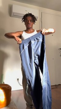 Moški v belem brezrokavniku drži modre športne hlače v notranjosti blizu belih vrat in klimatske naprave.