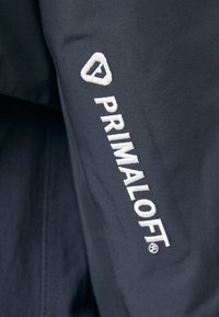 Dark fabric jacket featuring the "Primaloft" logo in white embroidery on the left sleeve. Smooth texture with a modern design.