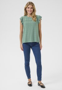 Top verde menta con maniche a volant, caratterizzato da motivi ad occhio di pernice. Abbinato a jeans skinny blu e ballerine nere con dettagli a staffa.
