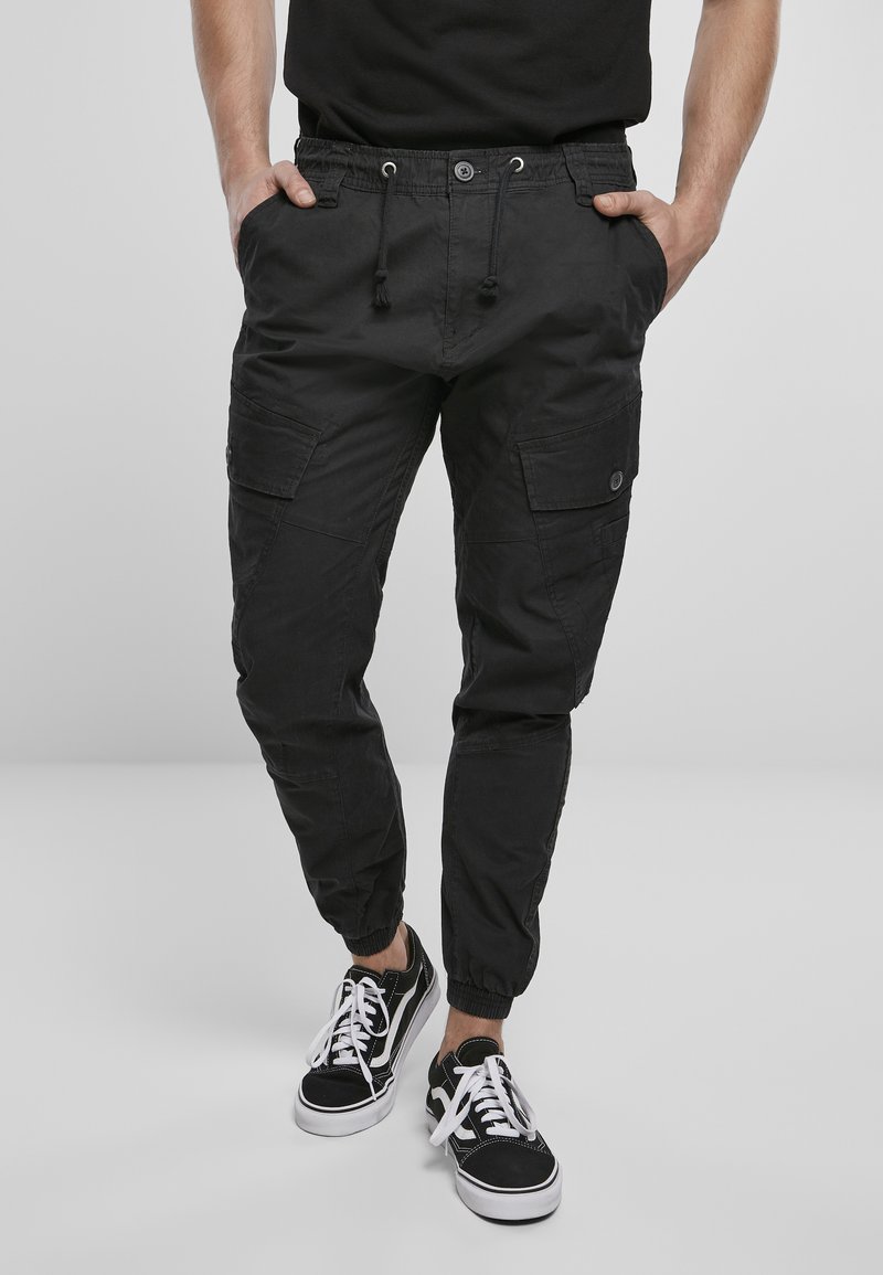 Pantalones cargo negros con puños elásticos, cintura con cordón y múltiples bolsillos laterales, combinados con zapatillas negras y cordones blancos.