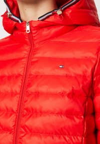 Chaqueta acolchada roja con diseño de acolchado horizontal, cierre de cremallera y capucha ajustable. Presenta un pequeño parche con el logotipo y accesorios metálicos brillantes.