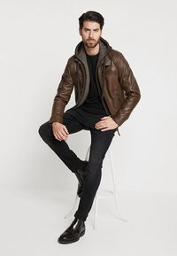 Bruine leren jas over een grijze hoodie, zwart t-shirt, donkere jeans en zwarte laarzen. Het model zit, met een ontspannen houding.