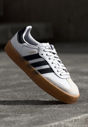 Witte Adidas Samba sneaker met zwarte strepen en een rubberen zool, licht gekanteld boven een getextureerd grijs oppervlak.