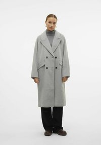 Cappotto grigio in misto lana con design doppiopetto, ampi revers, tasche laterali e maniche a vestibilità larga. Abbinato a pantaloni e scarpe nere.