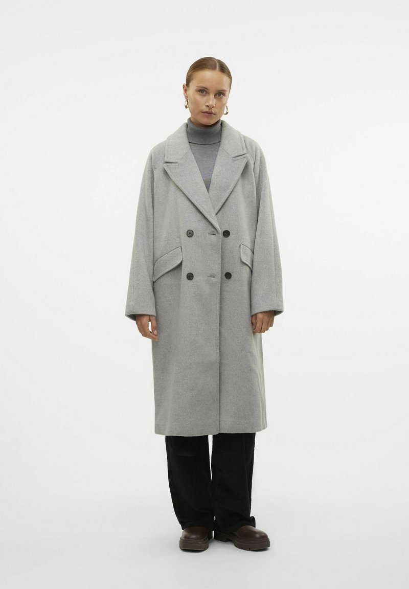 Cappotto grigio in misto lana con design doppiopetto, ampi revers, tasche laterali e maniche a vestibilità larga. Abbinato a pantaloni e scarpe nere.