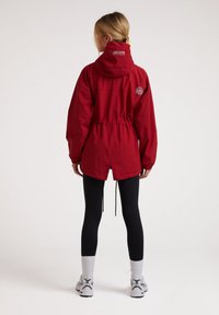 Rød letvægtsjakke med hætte, snoretræk i taljen og logo på ryggen. Båret over sorte leggings og hvide ankelsokker.