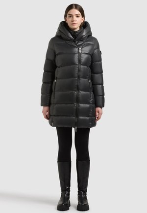 Mujer con un abrigo largo negro acolchonado con capucha, leggings negros y botas negras hasta la rodilla, de pie frente a un fondo claro y liso.