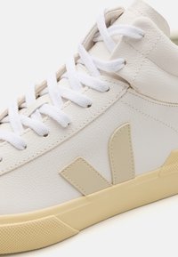 Bijela kožna tenisica s bijelim vezicama, teksturiranim jezikom i bež naglascima. Ima gumeni potplat s uzorkom za prianjanje.