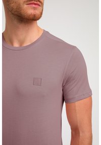 Kurzarm, helllila T-Shirt aus weichem Material, mit einem kleinen quadratischen Logo-Patch auf der Brust und einem runden Halsausschnitt.