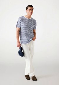 Tricou cu dungi albastre închis și albe, croială lejeră, asortat cu pantaloni bej deschis și pantofi maro slip-on. Ține în mână o șapcă albastru închis. Fundal simplu.