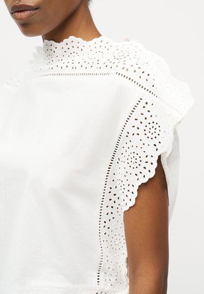 Κοντινό πλάνο ενός ατόμου που φοράει λευκό τοπ με διακοσμητικές λεπτομέρειες από δαντέλα με κυματιστό τελείωμα γύρω από το γιακά και τους ώμους, ενώ το χέρι είναι ακάλυπτο.