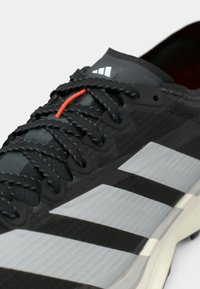 Μαύρο αθλητικό παπούτσι με υφασμένο επάνω μέρος, με γκρι ρίγες και μαύρα κορδόνια. Μια πορτοκαλί εσωτερική θηλιά βρίσκεται κοντά στη γλώσσα.