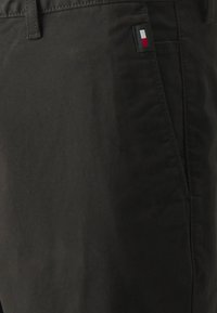 Pantaloni in cotone neri con un piccolo etichetta a bandiera sul lato, texture liscia e vestibilità sartoriale. Vista ravvicinata del tessuto.
