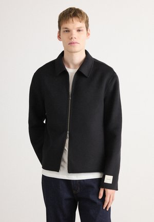 Black zipped jacket with a collar, made from a textured fabric. Features a straight cut and a small label on the left sleeve. Worn over a light shirt.