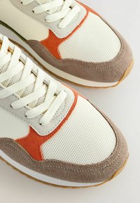 Baskets de sport avec un dessus en mesh crème, des accents en daim gris clair et orange, un talon en daim marron texturé et une semelle en caoutchouc blanche.