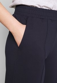 Hand in de zak van marineblauwe elastische taillebroek, gedragen met een lichtgekleurde ingestopte blouse.